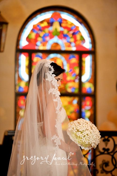 A bride in front of stained glass window holding her bouquet.