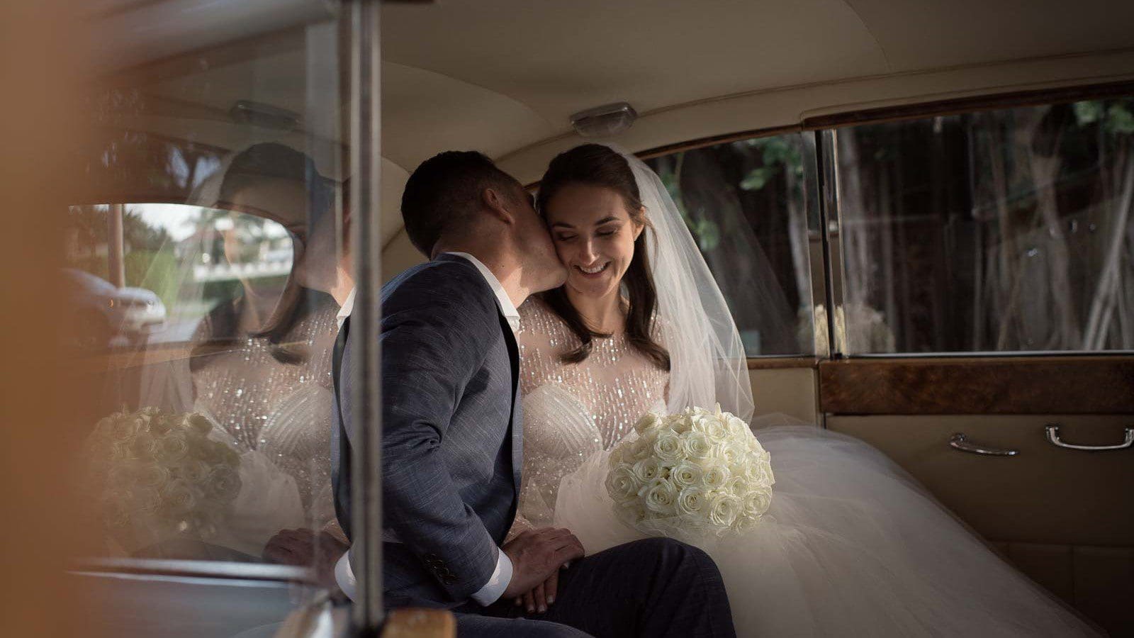 A bride and groom sharing a tender moment inside a vintage car, with the bride holding a bouquet and both partly obscured by her veil.