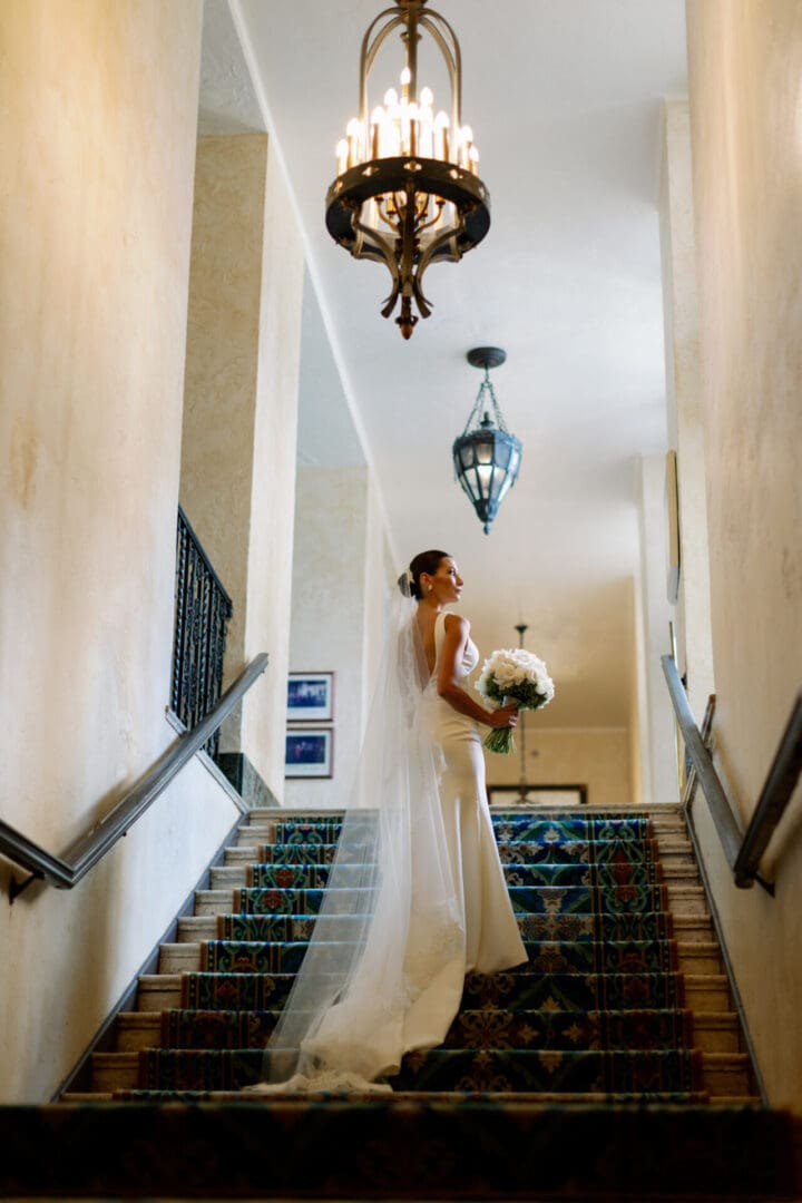 A bride in a sleek white dress and long veil ascends a staircase adorned with colorful tiles, holding a bouquet, under elegant hanging chandeliers.