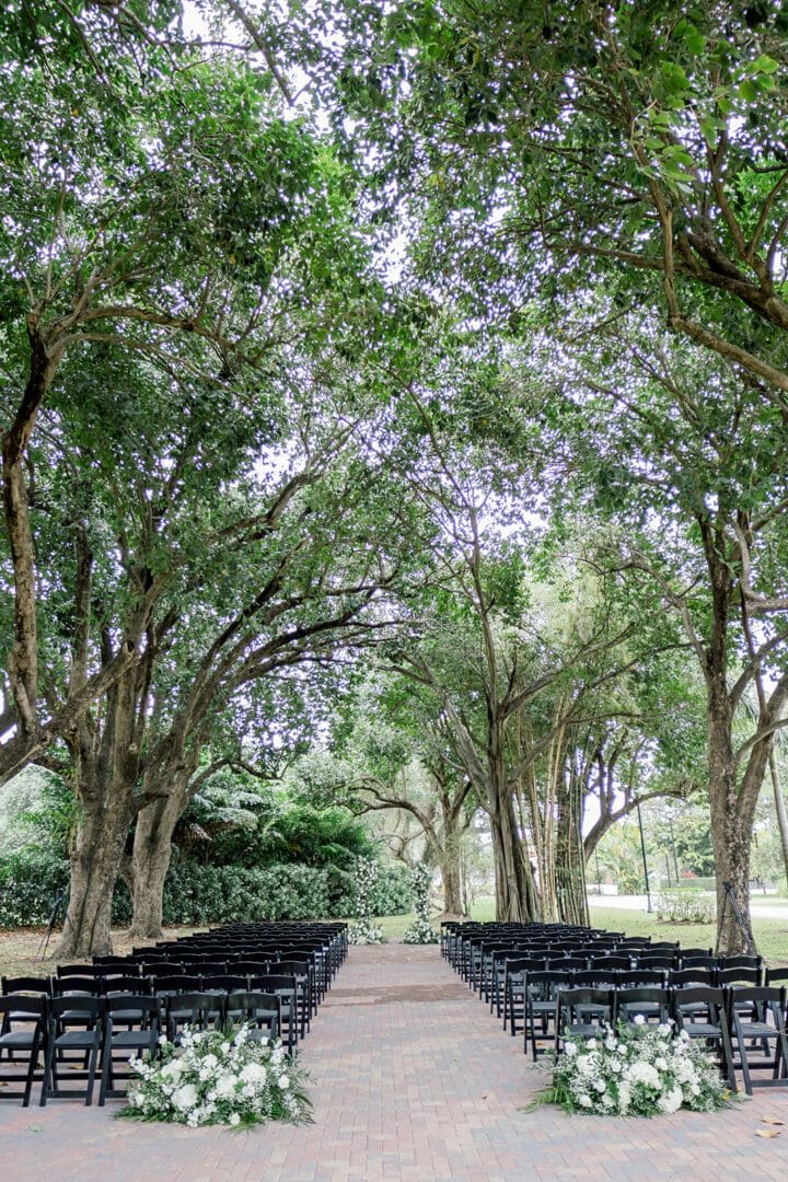 Outdoor wedding venue setting with rows of black chairs and floral decorations along a central aisle, surrounded by large, leafy trees.