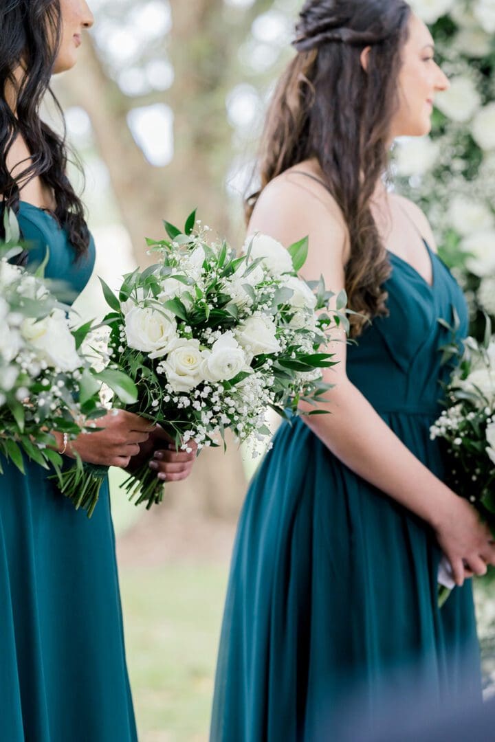 Two women in teal dresses holding large white floral bouquets at an outdoor setting, partially obscured by green foliage.