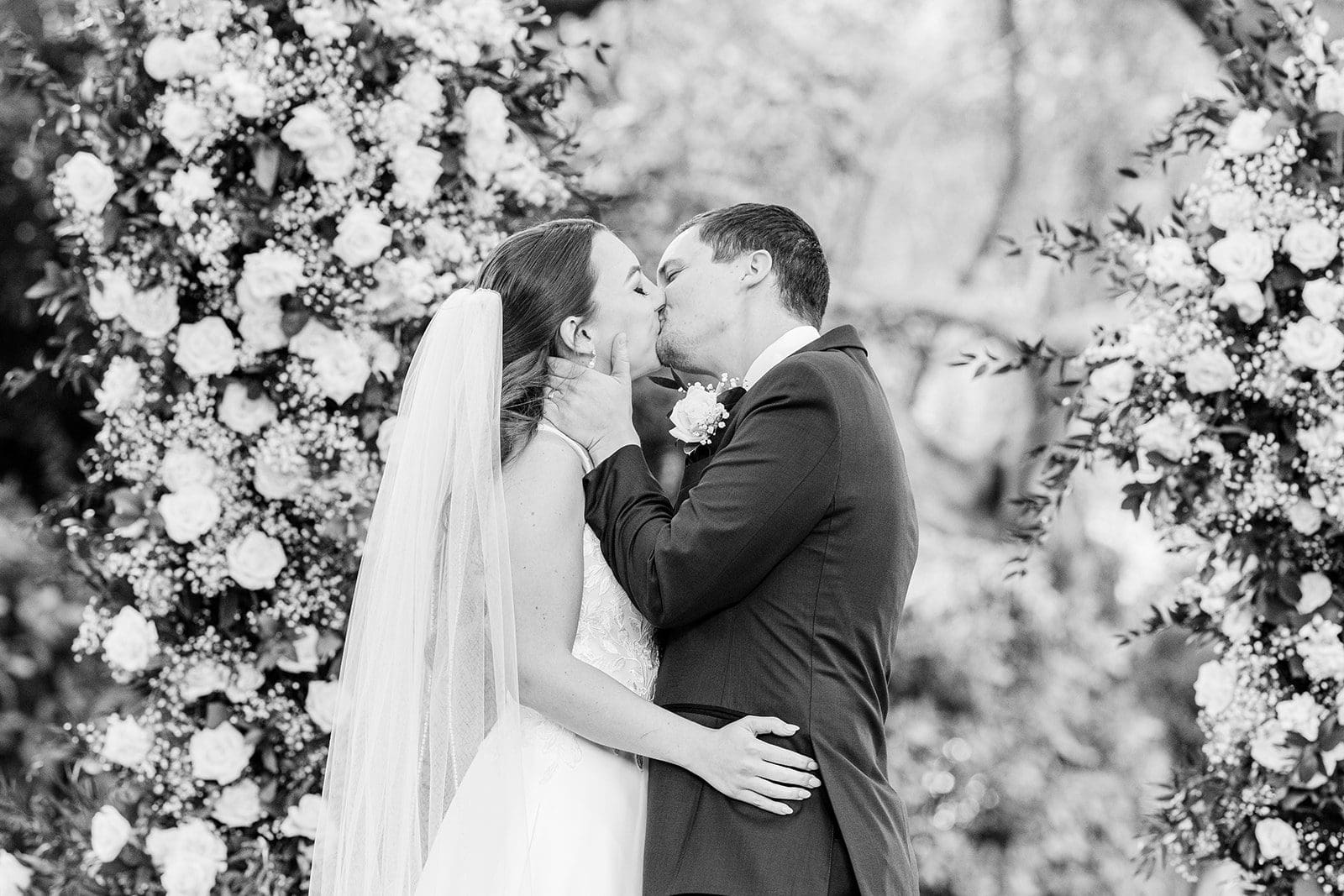 A bride and groom kissing, surrounded by lush white flowers. the image is in black and white.