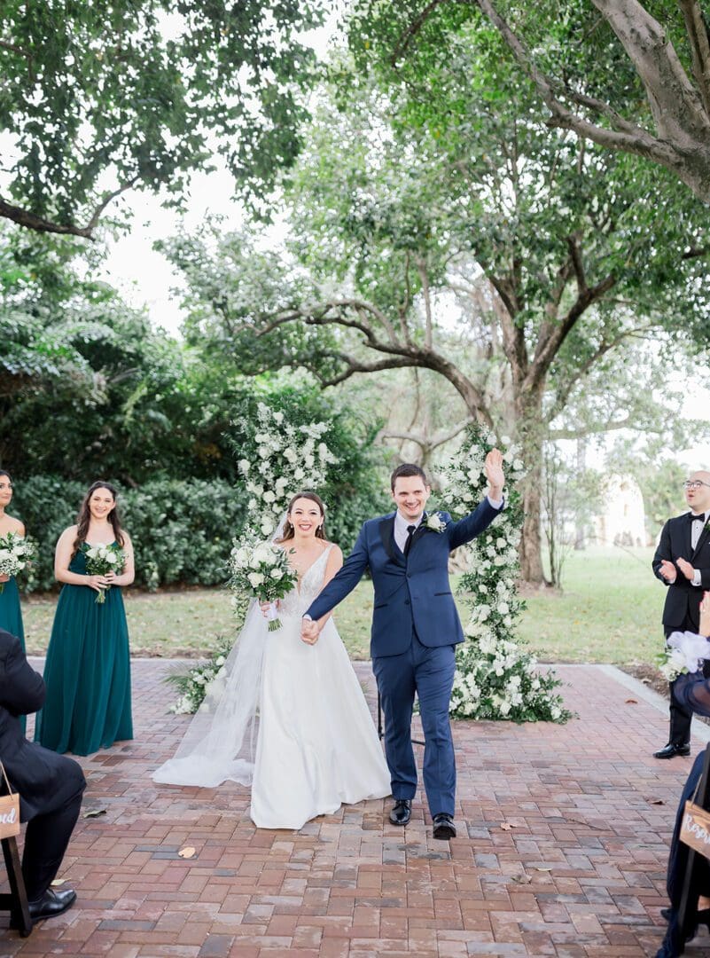 A couple is waving at the camera while standing under trees.