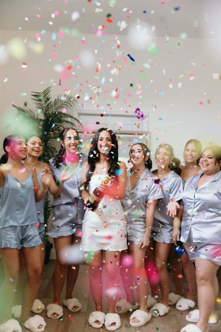 A group of women in party attire smiling and celebrating with confetti in the air.