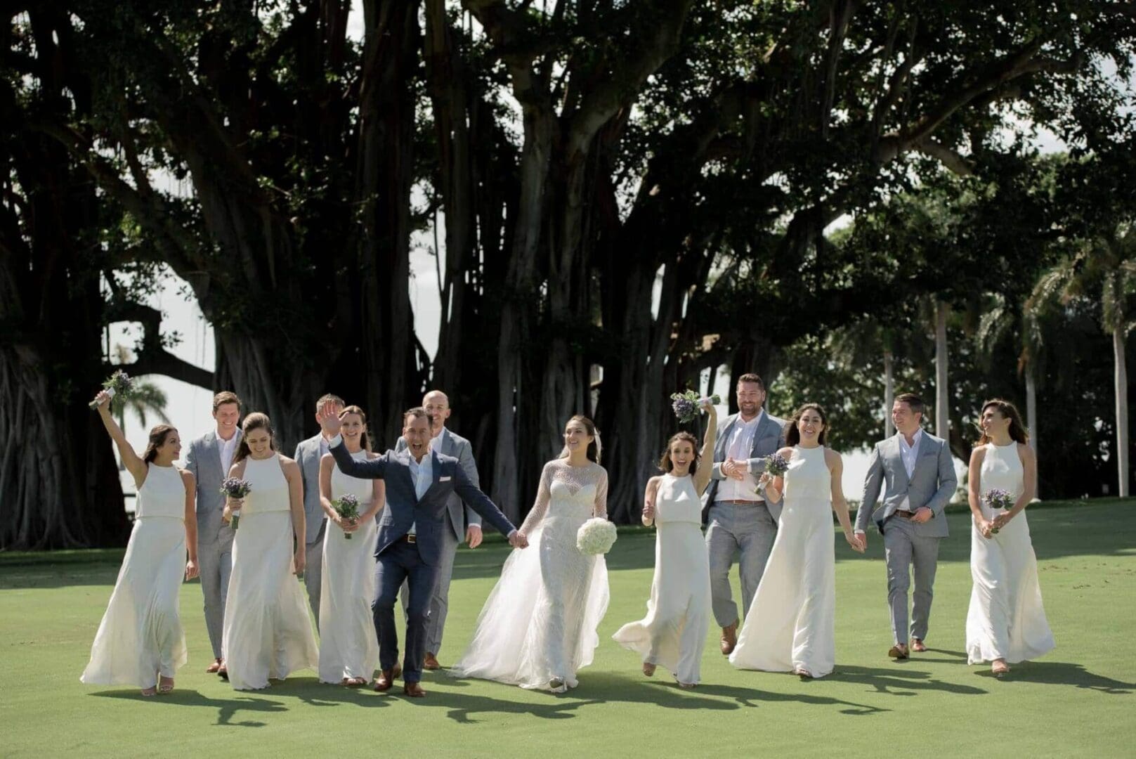 A wedding party walking joyfully on a grassy lawn under large trees, with the bride and groom leading, surrounded by bridesmaids and groomsmen.