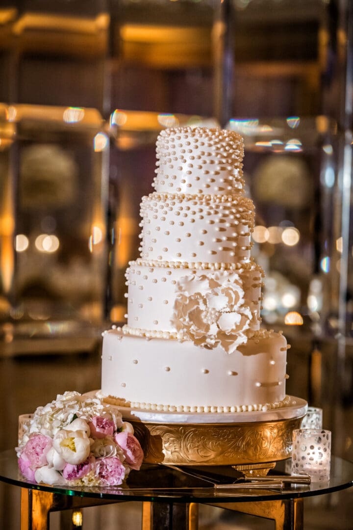 Elegant multi-tiered wedding cake decorated with pearls and floral accents, displayed on a golden tray against a softly lit, reflective background.