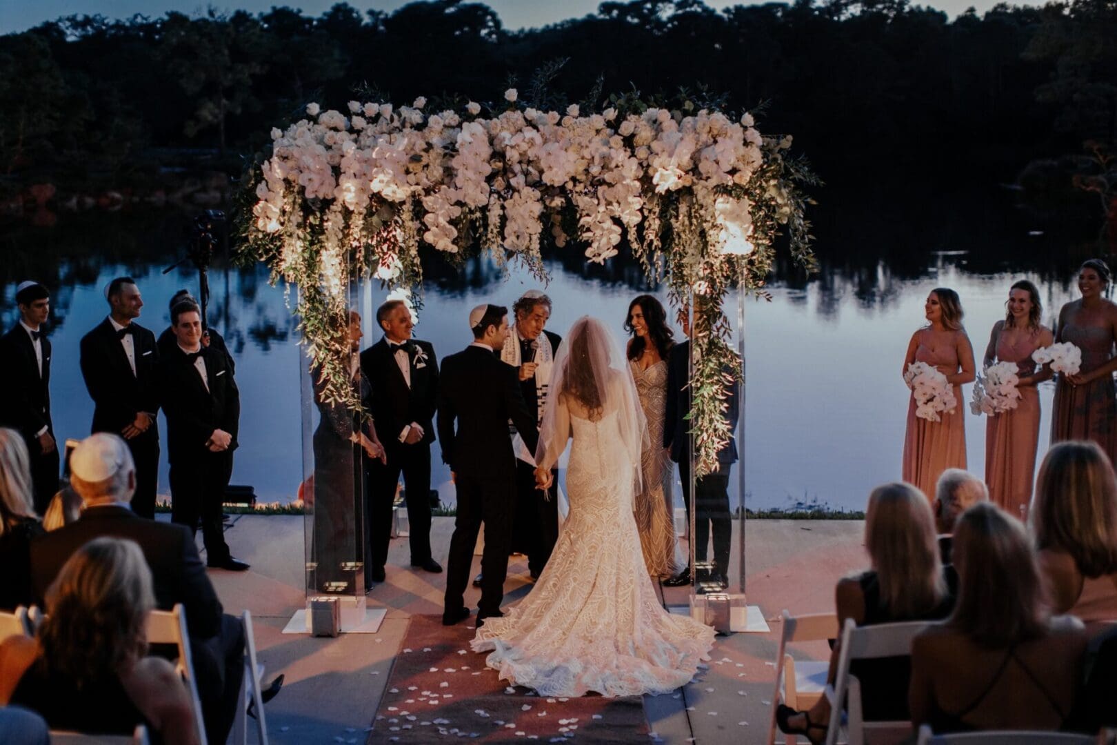 A wedding ceremony by a lake at sunset, featuring a couple at the altar, surrounded by attendants and guests, with floral decorations.