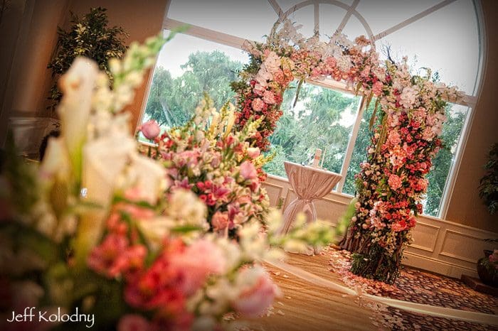 Elegant wedding arch decorated with pink and white flowers inside a room with a window view of greenery, with a small table and chairs nearby.