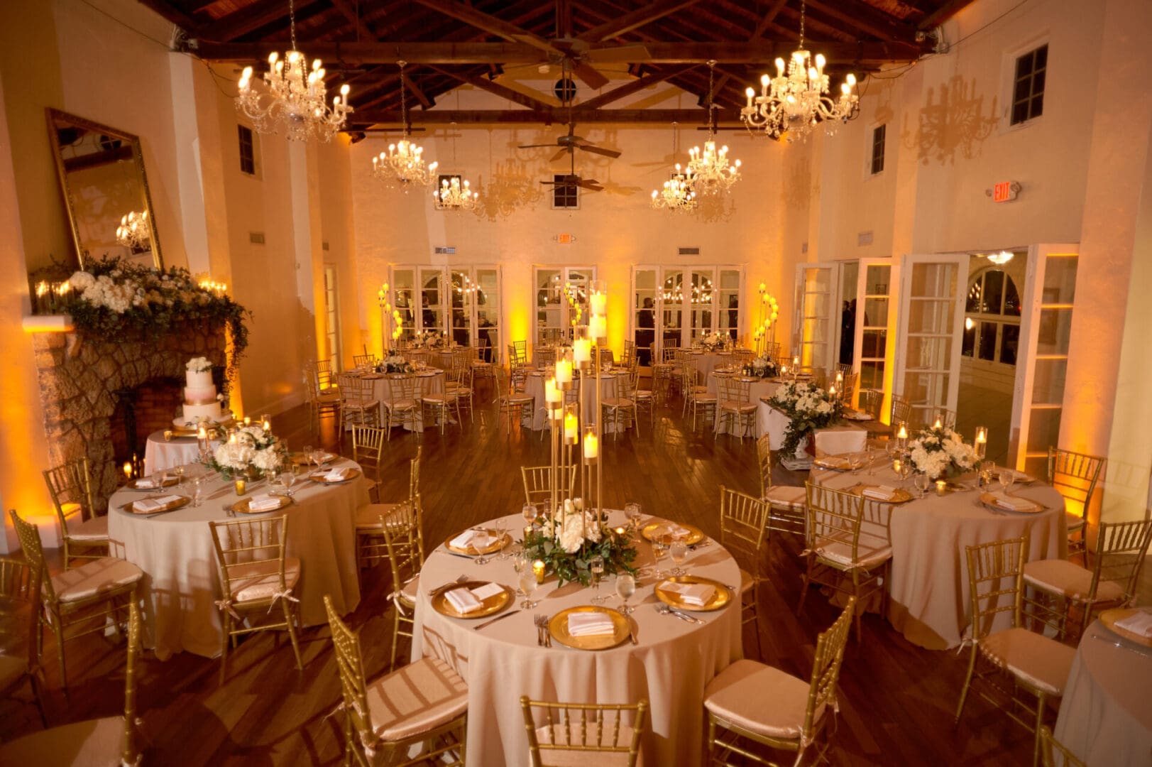 Elegant wedding reception hall with round tables set for dinner, floral centerpieces, and chandeliers hanging from a wood-beamed ceiling, warmly lit.