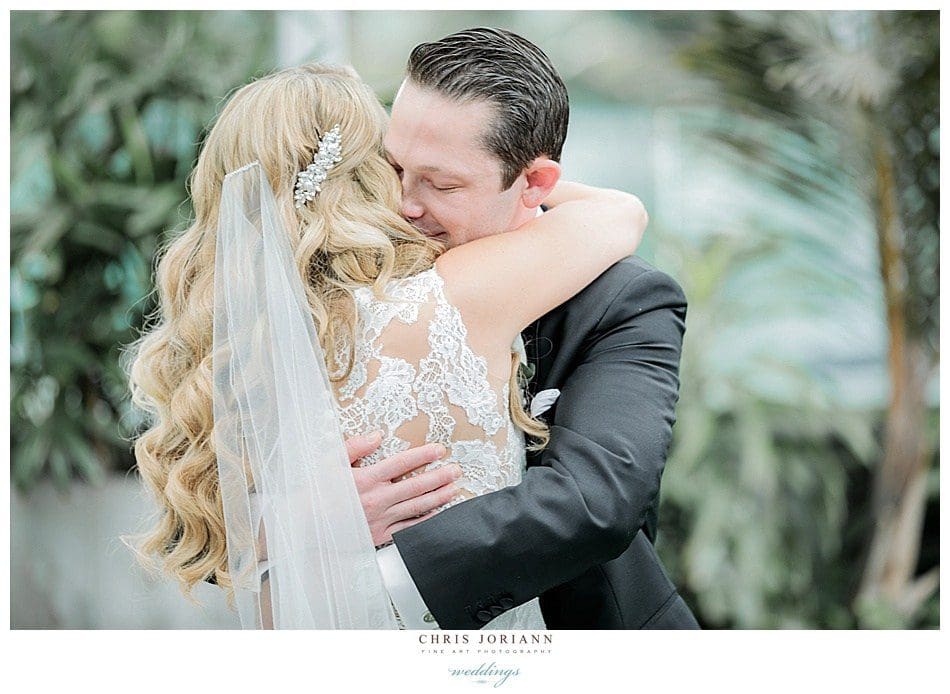 A bride and groom embrace and kiss, surrounded by greenery. the bride's long, curly hair and detailed lace dress are prominent.