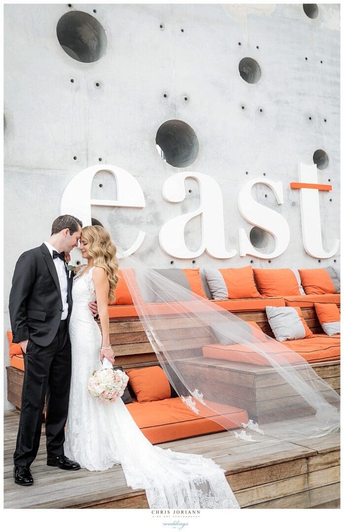 A wedding couple sharing a kiss in front of a modern wall with climbing holds and a neon "east" sign, with orange seat cushions and wooden slats in the background.