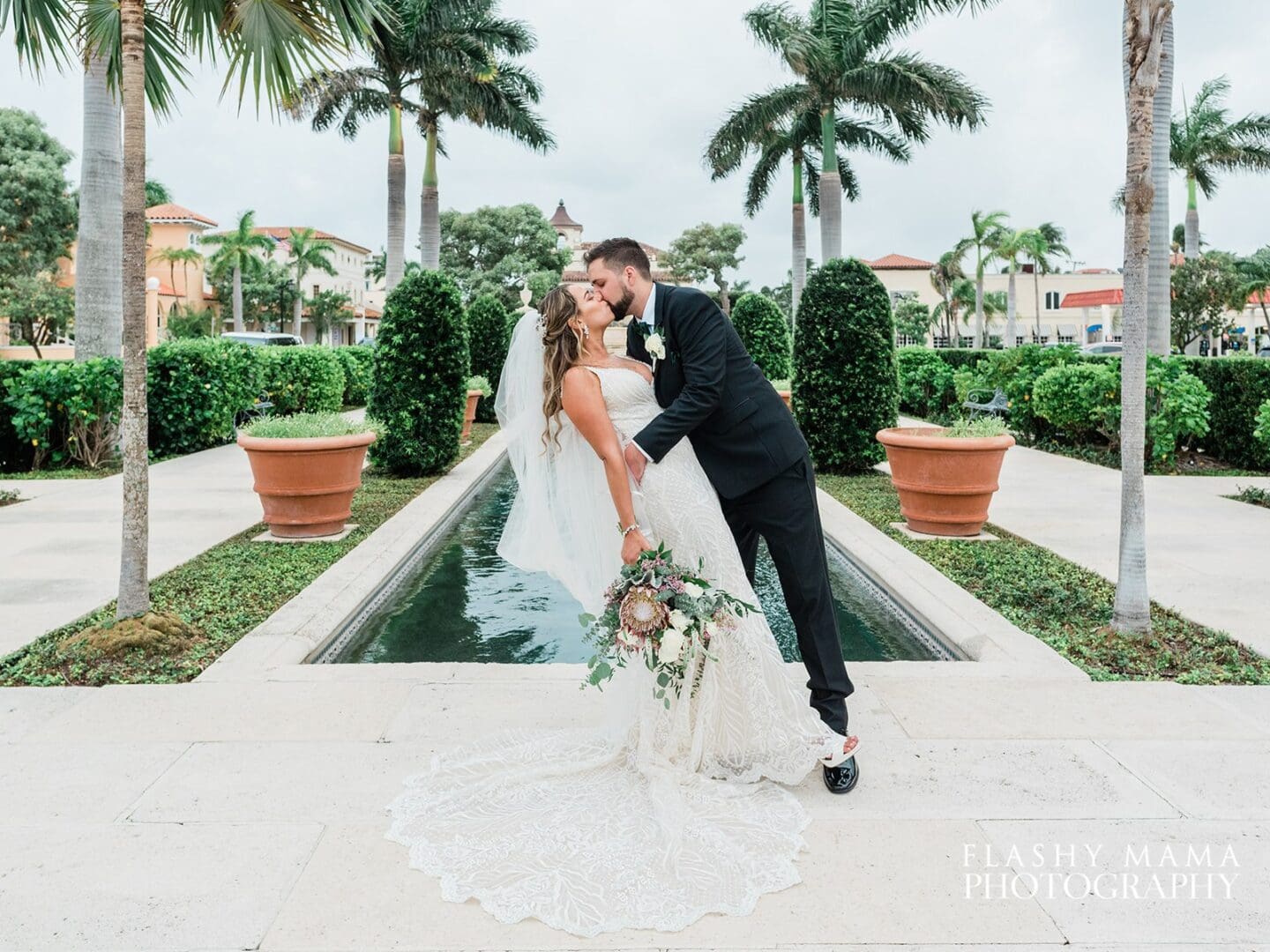 A bride and groom kiss romantically beside a narrow outdoor water feature, surrounded by palm trees and manicured shrubs, under an overcast sky.