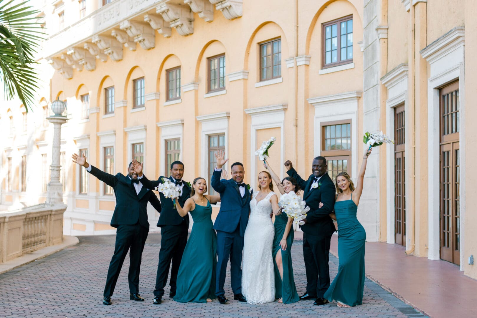A joyful wedding group, including the bride and groom, celebrates outside a grand, peach-colored building, raising bouquets and arms in excitement.