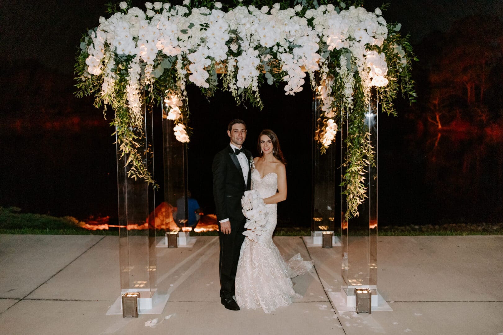A bride and groom standing under a floral arch at night with candles lining the pathway.