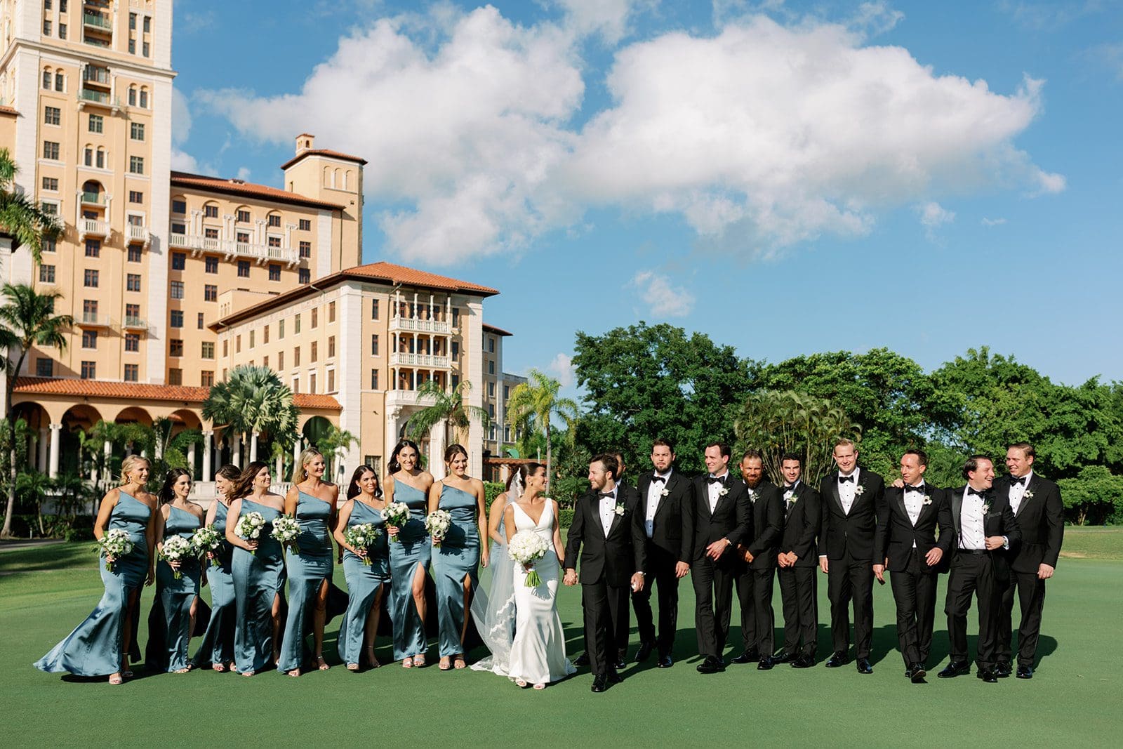 A wedding party, with bridesmaids in blue dresses and groomsmen in black tuxedos, stands on a green lawn before a large hotel building.