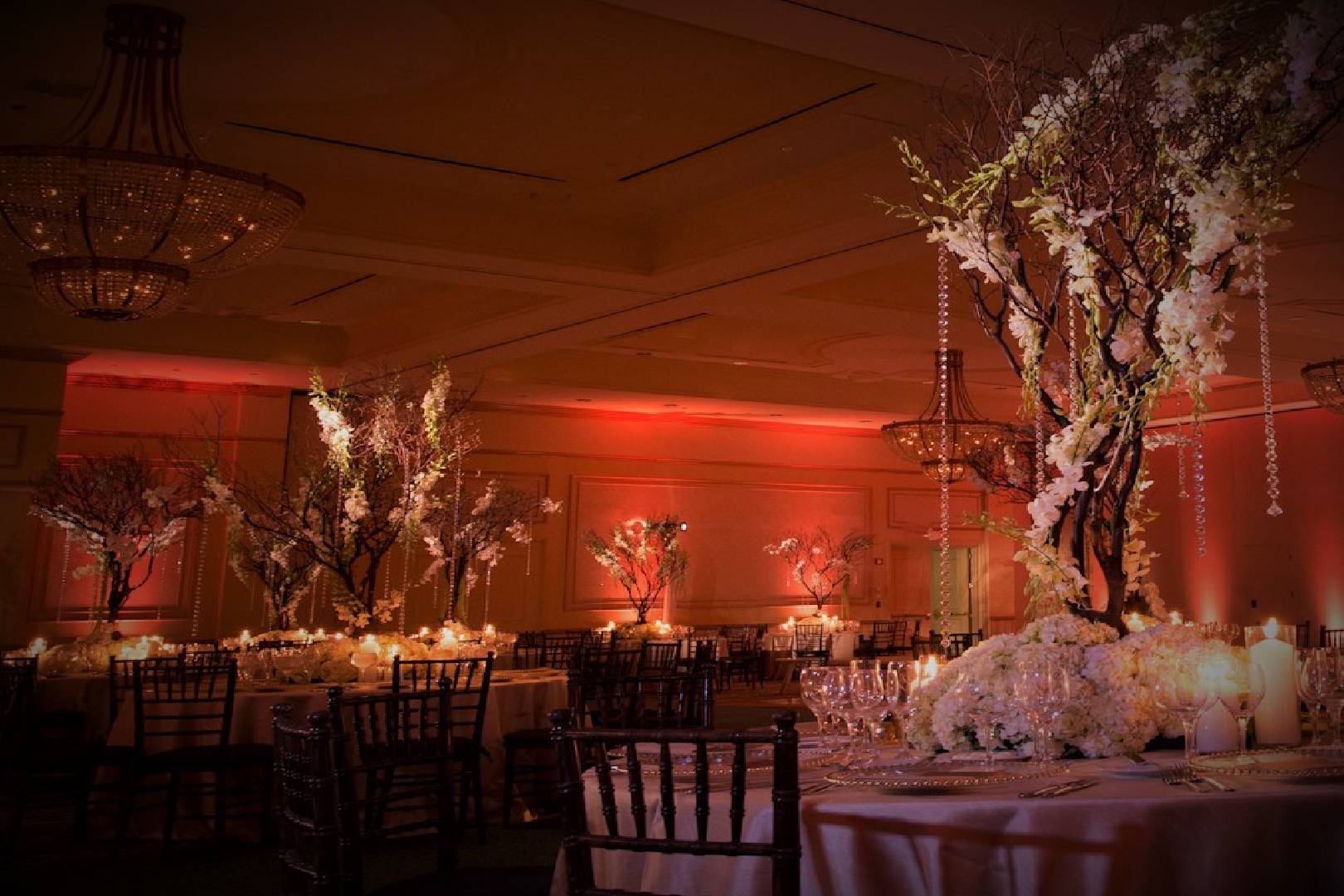 Elegant banquet hall decorated with floral centerpieces, lit candles on tables, and ambient red lighting, with grand chandeliers overhead.