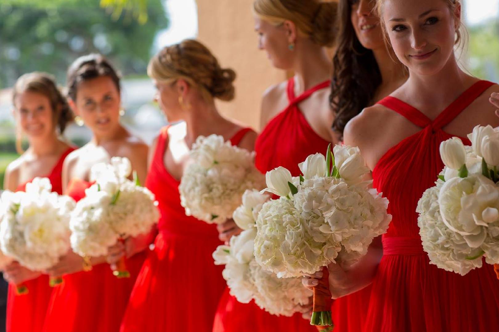 Five bridesmaids in red dresses holding white bouquets, standing in a line, with one looking at the camera.