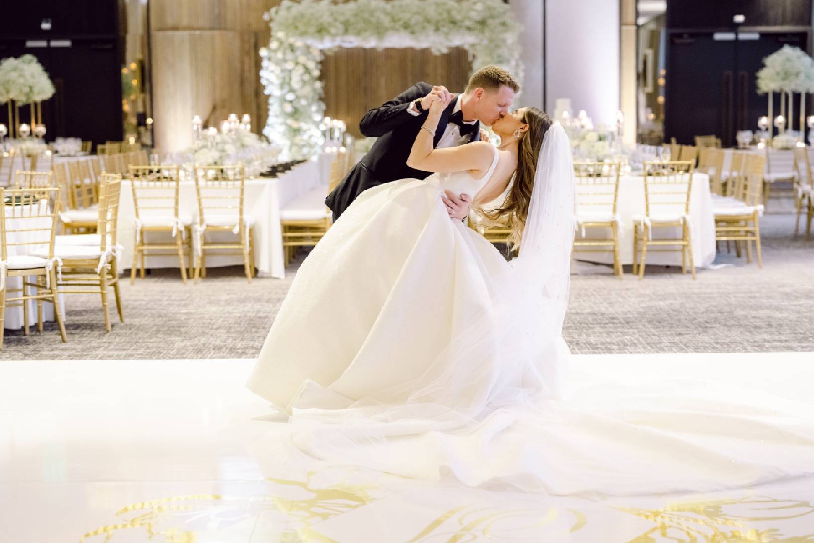 A bride and groom share a kiss in a lavishly decorated ballroom with elegant chairs and floral arrangements in the background.