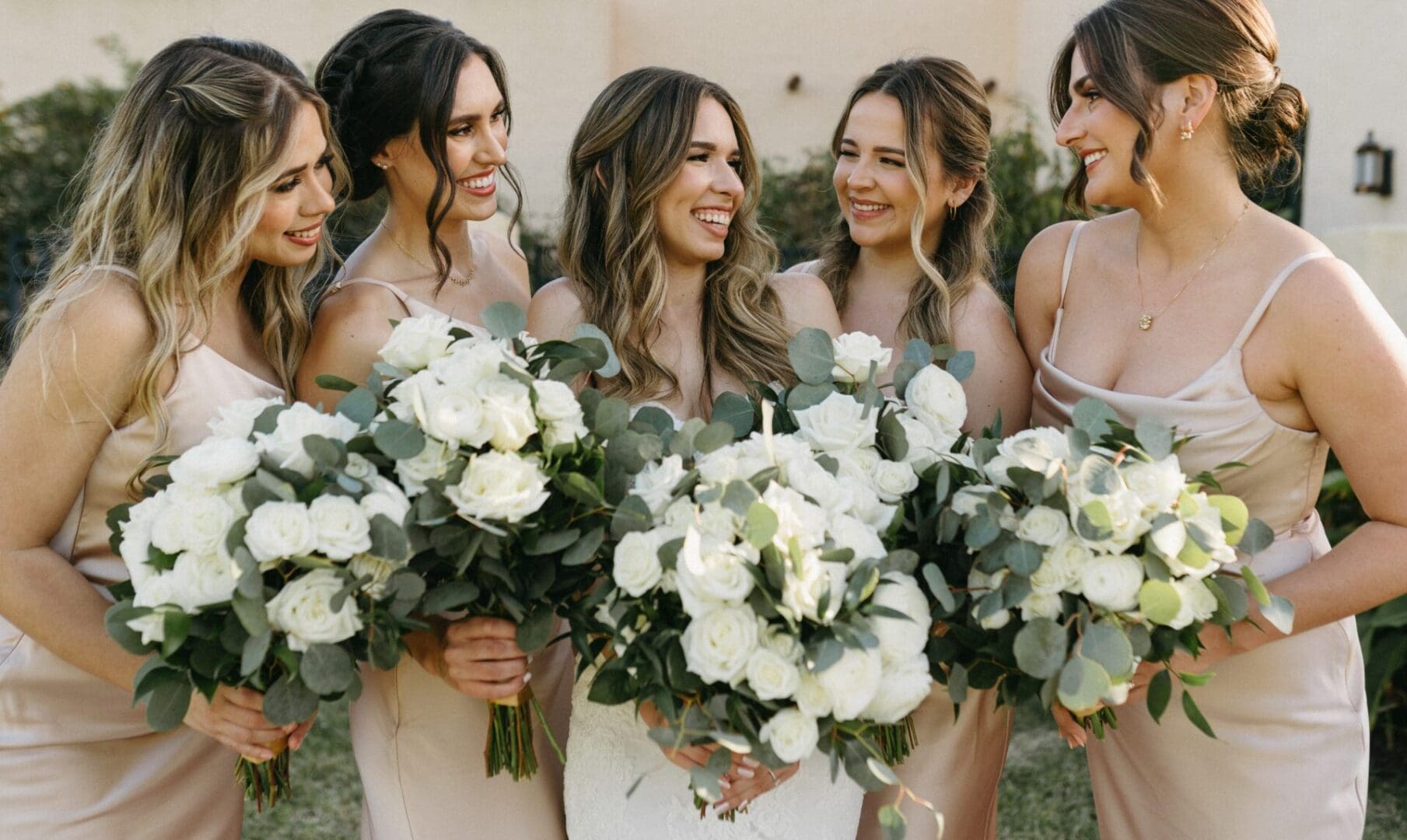 Five women in pastel dresses holding white bouquets and smiling at a wedding event.