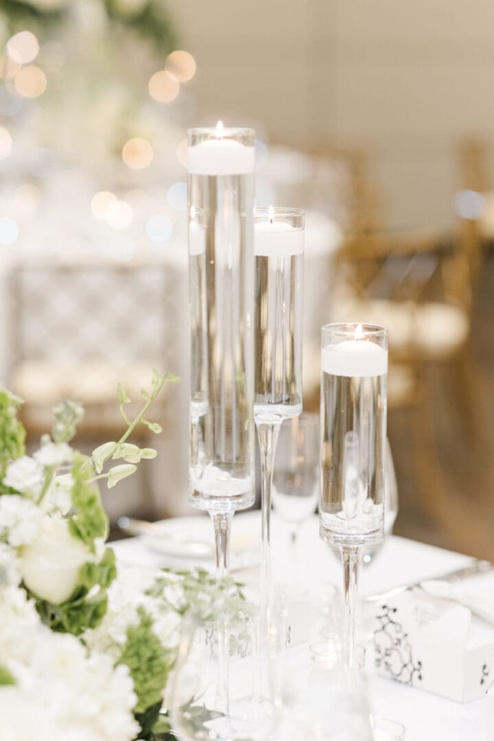 Elegant wedding table setting featuring tall glass candleholders with lit candles, surrounded by white floral arrangements and soft lighting.