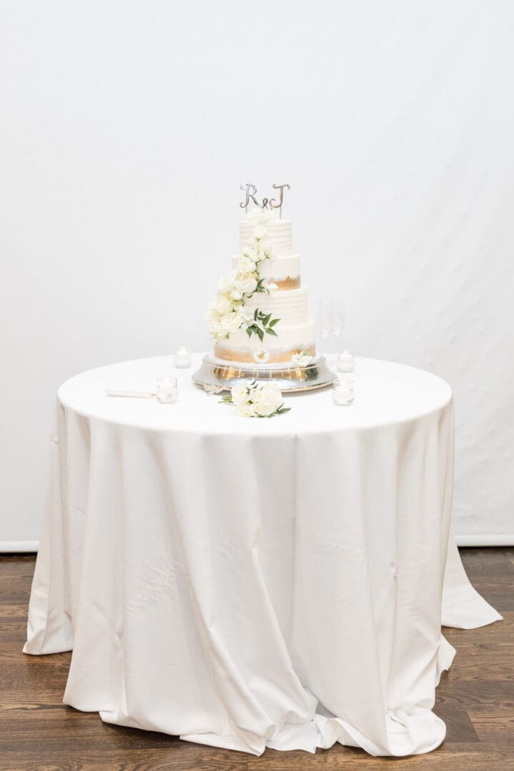 An elegantly decorated wedding cake on a round table draped with a white cloth, adorned with white flowers and initials on top.