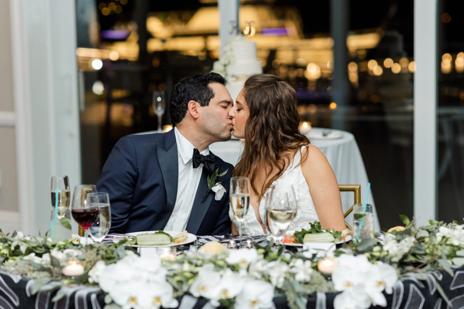 A bride and groom kissing at a wedding reception table, decorated with white flowers and candles, with a cake in the background.