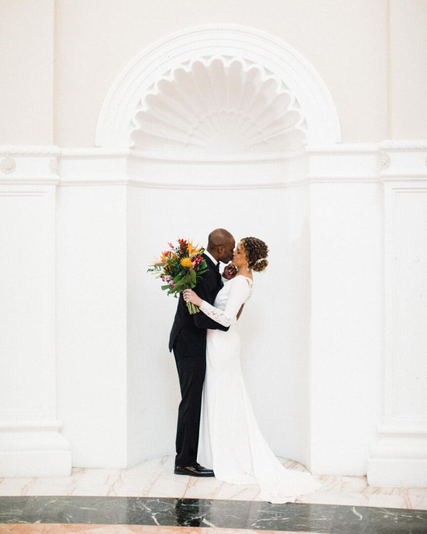 A bride and groom embracing and kissing, the bride holding a colorful bouquet, in front of a white arched backdrop.
